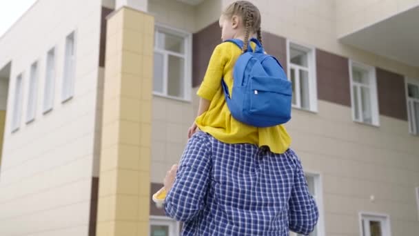 Little daughter sits on the shoulders of dad, they go to school together. Preschool education. Happy family, father and child go to school. Baby, goes to study. Family outing after school — Stock Video