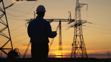 Beyaz miğferli Siluet Güç Mühendisi güç hattını kontrol ediyor tabletteki bilgisayarı kullanarak, güç sisteminin uzaktan kumandasıyla. Gün batımında yüksek voltajlı elektrik hatları. Elektrik dağıtımı ve tedariği