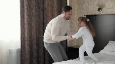 Mutlu aile çocuğu, kız ve baba, el ele tutuşup yatağa atlıyorlar. Babası neşeli bir şekilde gülüyor. Sevimli kızıyla aktif bir oyun oynuyor. Yatak odasında bir çocuk. Sabahları dans ediyorum. Baba izin günü.