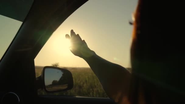 Девушка с длинными волосами сидит на переднем сиденье автомобиля, вытягивает руку из окна и ловит сияние заходящего солнца. Бесплатная женщина путешествует на машине ловит ветер рукой из окна машины. Отпуск — стоковое видео