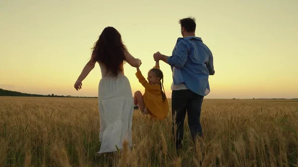 Barnet leker, hoppar, springer bredvid mamma, pappa håller hand på fältet. Lycklig familjevandring på ett vetefält vid solnedgången. En bondefamilj. Föräldrar, kära baby, går glatt tillsammans i solens strålar. — Stockfoto