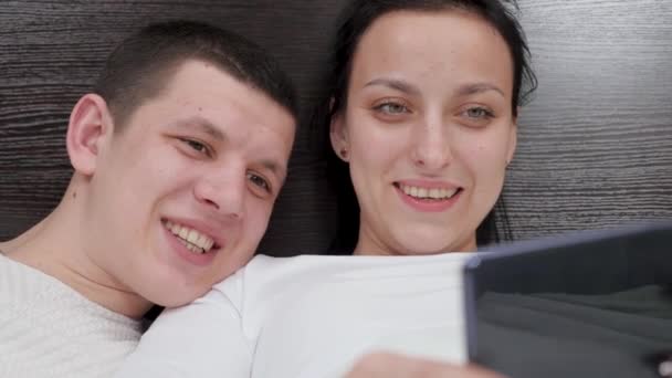 Una joven pareja que sostiene un teléfono inteligente en la mano es fotografiado juntos, dispara video blog en la cama. Feliz marido y mujer de la familia tomar fotos en línea utilizando un teléfono móvil moderno en casa en el sofá en el dormitorio. — Vídeos de Stock