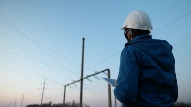 Beyaz kasklı kadın güç mühendisi güç hattını kontrol ediyor tabletteki bilgisayarla, güç sisteminin uzaktan kumandasıyla. Gün batımında yüksek voltajlı elektrik hatları. Dağıtım ve elektrik tedariği. Enerji