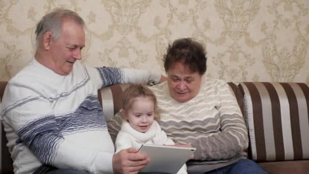 Moderne grootouders spelen met kind, meisje zit met digitale tablet online thuis. Gelukkige familie. Een ouder stel met kleindochter. Kind toont iets in tablet aan zijn grootouders. — Stockvideo