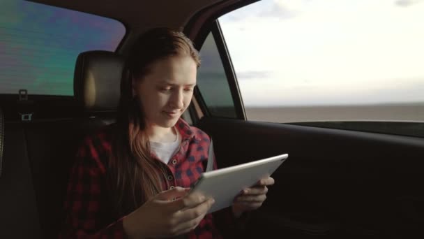 Estudante estuda, trabalha remotamente. Menina bonita passageiro viaja de carro com tablet computador, sentado no banco de trás do carro e olha para a janela aberta. Mulher livre está desfrutando de passeio de carro e descanso. — Vídeo de Stock