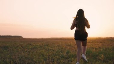 Güneşin peşinden koşmak. Antrenman koşusu. Sağlıklı, güzel bir kız spor yapıyor, kırsalda güneş altında koşuyor. Koşucu kız sahada temiz hava soluyor. Özgür genç kadın gün batımında yaz parkında koşuyor..