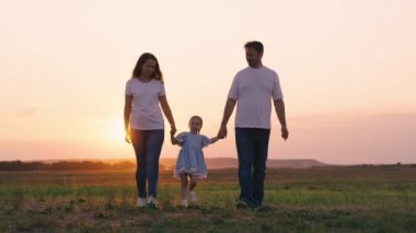 Anne, baba ve kızı yürüyorlar, ebeveynlerinin ellerini tutuyorlar, çocuk yeşil çimlerin üzerinde zıplıyor. Mutlu aile baharda gün batımında parkta yürür. Mutlu sağlıklı bir çocukluk. Aile hafta sonu