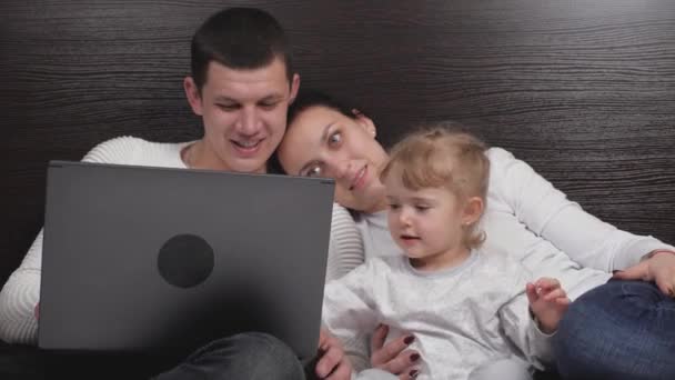 En glad familie med et lille barn studerer online på computeren derhjemme, barn ønsker ikke at studere, leger med sine forældre. Datter ser tegneserie med mor og far ved hjælp af bærbar computer, mens du sidder på sengen – Stock-video