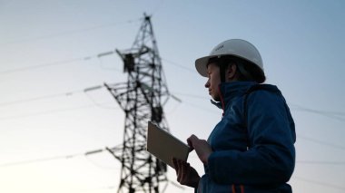 Beyaz kasklı bir kadın enerji işçisi tabletteki bir bilgisayarla güç sistemini kontrol ediyor. Yüksek voltajlı elektrik hatları. Çevre dostu elektrik enerjisi