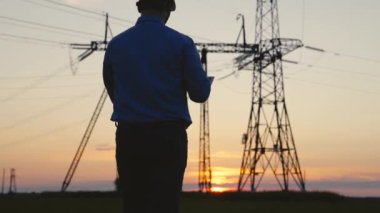 Beyaz miğferli Siluet Güç Mühendisi güç hattını kontrol ediyor tabletteki bilgisayarı kullanarak, güç sisteminin uzaktan kumandasıyla. Gün batımında yüksek voltajlı elektrik hatları. Elektrik dağıtımı ve tedariği