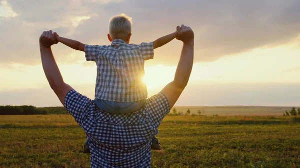 一个快乐的孩子，一个儿子坐在他父亲的脖子上，扮演一个飞行员，像一个超级英雄一样飞翔，梦想着和他的父亲一起飞翔。爸爸和孩子的游戏，幻想。快乐的一家人在阳光下在公园里休息. — 图库照片
