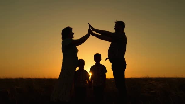 Feliz família sonha com sua própria casa no parque no verão ao sol. Crianças mamãe e papai estão brincando juntos construindo casa com as mãos, silhueta ao pôr do sol. Cuidar das crianças pelos pais — Vídeo de Stock