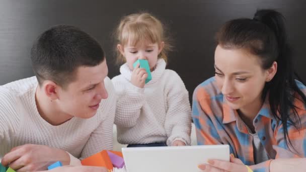 現代のガジェット、親と子供は一緒にデジタル教育の概念を勉強します。幸せな家族、両親と小さな子供、娘一緒に、ベッドの上に横たわってタブレットを使用します。母と子と父が家で — ストック動画