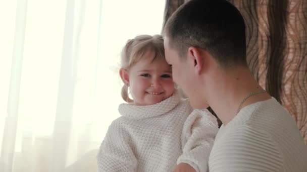 Папа играет со своей маленькой дочерью в комнате, девочка обнимает своего любимого папу у окна. Счастливая семья, молодой отец, играет с маленьким очаровательным ребенком, дочь весело улыбается на руках отца — стоковое видео