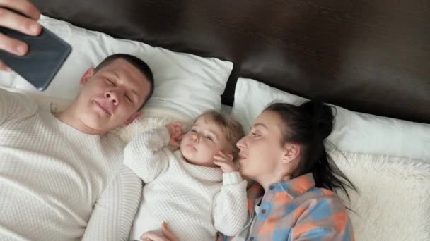 Felice giovane famiglia con una bambina, sono fotografati su uno smartphone nel letto. Madre, padre e figlia-blogger in camera comunicano utilizzando un moderno gadget online. Moderna giovane famiglia — Video Stock