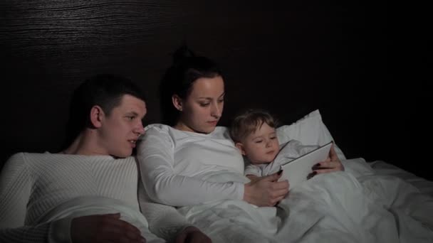 Educación familiar en línea de los niños. Familia feliz descansando juntos en la noche en casa con una tableta. Niño pequeño, hija viendo dibujos animados en línea usando una tableta digital, sentado en la cama. — Vídeo de stock