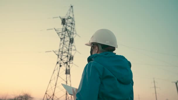 Uma mulher trabalhador de energia em um capacete branco funciona verifica a linha de energia usando um computador em um tablet, controla o sistema de energia. Linhas eléctricas de alta tensão. Energia elétrica ecológica — Vídeo de Stock