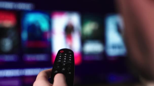 Un hombre mira televisión inteligente y usa control remoto negro. Las páginas borrosas de los rollos de televisión. Mans mano selecciona los canales de televisión de Internet con control remoto, primer plano. La persona controla la televisión usando un control remoto moderno. — Vídeo de stock