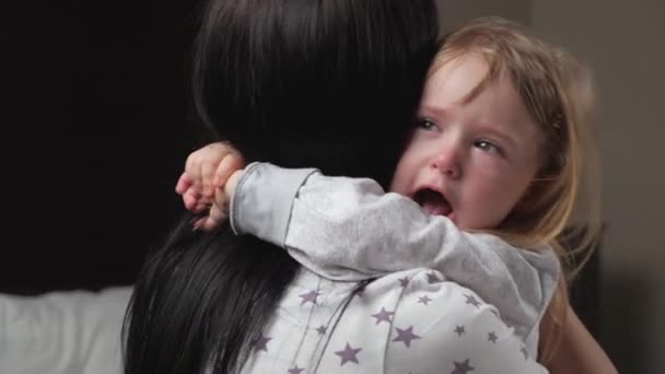 Мама заспокоює свою дочку, дитина плаче в кімнаті на руках своєї матері. Любляча молода мати обіймає і втішає свою маленьку дочку. Родина, дитина зі сльозами на очах обіймає матір. — стокове відео