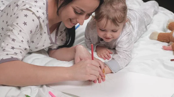 Mom, nanny, teaches girl to draw, palm is outlined with pencil. Happy family playing together at home on bed. Mom helps her daughter learn to draw on paper, coloring with multi-colored pencils — Stock Photo, Image