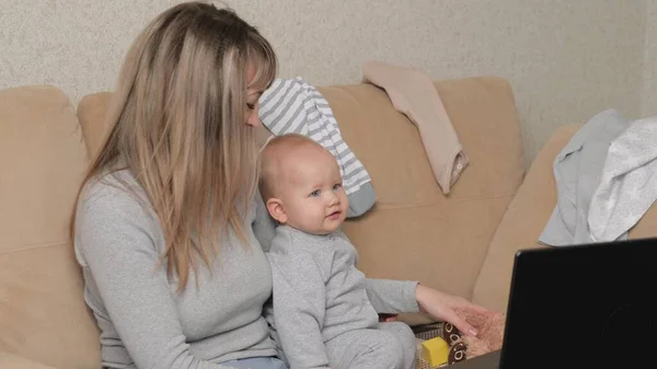 Mãe e bebê estão assistindo a um desenho animado em um computador, o bebê sorri. Babá com um lindo bebê em seus braços em uma casa acolhedora. Trabalho freelancer feminino. Maternidade moderna, multitarefa — Fotografia de Stock