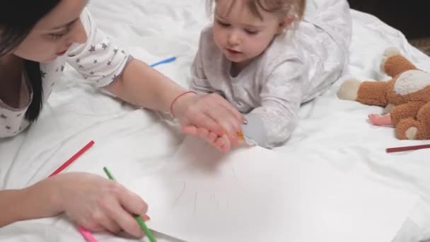 Mamma, tata, insegna alla ragazza a disegnare, il palmo della mano è delineato con la matita. Famiglia felice che gioca insieme a casa a letto. La mamma aiuta sua figlia a imparare a disegnare sulla carta, colorando con matite multicolori — Video Stock