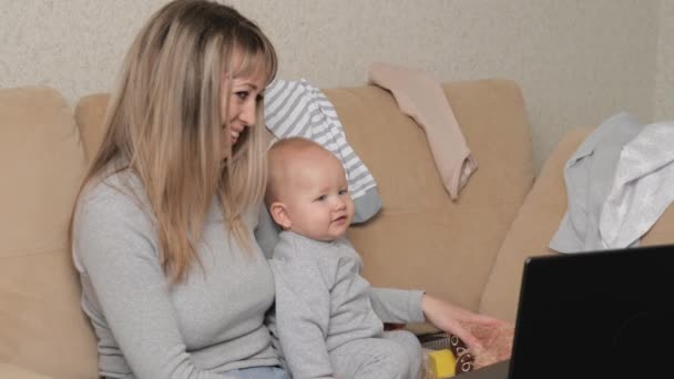 Mutter und Baby schauen sich am Computer einen Zeichentrickfilm an, das Baby lächelt. Kindermädchen mit einem wunderschönen Baby auf dem Arm in einem gemütlichen Zuhause. Frauen arbeiten freiberuflich. Moderne Mutterschaft, Multitasking — Stockvideo