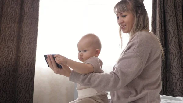 Счастливая семья, мать с маленьким ребенком, используют современный смартфон дома. Концепция семьи, технологии, материнства и людей. Видеозвонок папе, маленькой дочери и маме. Современное домашнее образование — стоковое фото