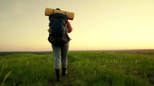 Una joven caucásica activa con mochila va de excursión. Mujer viajera libre caminando por el prado en verano. Chica viajero viaja a lo largo de la cima de la colina verde en el sol, meditación, ecoturismo, caminata —  Fotos de Stock