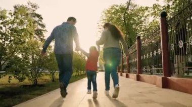 Anne, baba, kız şehir parkında birlikte oynuyorlar. Mutlu çocuk, atlayan ebeveynlerin elini tutuyor. Mutlu bir aile el ele tutuşuyor, ilkbaharda, yazları sokakta koşuyor. Mutlu sağlıklı bir çocukluk. Takım çalışması.