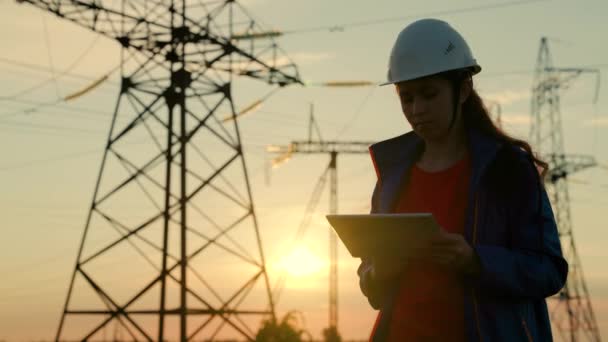 Ženský inženýr v bezpečnostní helmě kontroluje elektrické vedení pomocí digitálního tabletu. Elektrárenská společnost zaměstnankyně pracující venku, obsluha vysokonapěťového vedení při západu slunce — Stock video