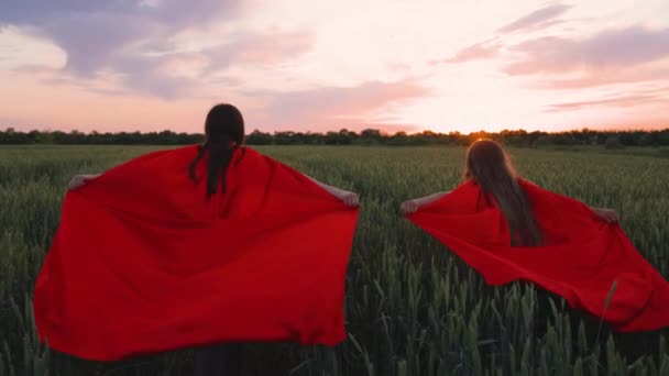 Barn, tonåringar och drömmar om att bli superhjälte. Unga flickor i röd mantel. Glada flickor spelar superhjältar, de springer över gröna fältet i röda mantlar, kamouflerar fladdrar i vinden. Barndrömmar — Stockvideo