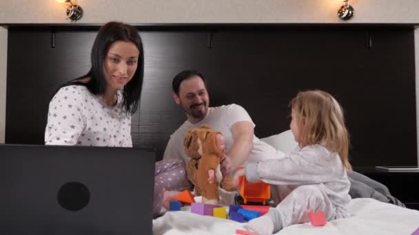 Μια νεαρή μητέρα εργάζεται στο σπίτι χρησιμοποιώντας ένα φορητό υπολογιστή, το βράδυ στο κρεβάτι το παιδί και ο μπαμπάς παίζουν παιχνίδια μαζί. Ευτυχισμένη οικογένεια στο σπίτι. Κόρη και ο μπαμπάς παίζουν με αρκουδάκι και αυτοκίνητα, μαμά μαθαίνει εξ αποστάσεως σε απευθείας σύνδεση. — Αρχείο Βίντεο