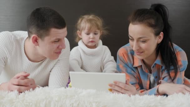 Gadget moderne, les parents et les enfants regardent dessin animé. Famille heureuse, parents et petit enfant, fille utilisent la tablette ensemble, couché sur le lit. Mère, enfant et père se reposent le week-end ensemble à la maison — Video