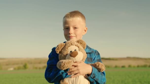 A criança brinca com o urso Teddy. Menino abraça seu brinquedo de urso macio favorito no playground. Brinquedo de pelúcia nas mãos de criança no parque de verão. O miúdo brinca com um brinquedo e sonha ao ar livre. Melhores amigos — Vídeo de Stock