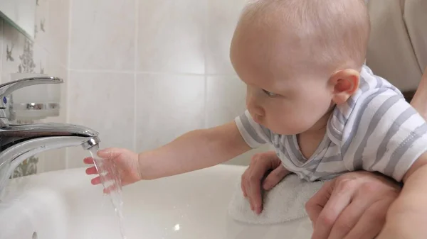 O miúdo aprende a lavar as mãos sozinho. Uma criança pequena lava as mãos com a mãe no banheiro acima da pia. Uma mãe ajuda seu filho a lavar as mãos em casa. Uma família feliz. Jogar água com criança. — Fotografia de Stock