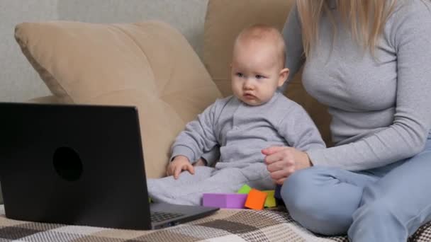 La maternidad moderna. Mamá le está enseñando a su hijo pequeño usando la computadora. Mujer ocupada trabajando en el portátil con su hijo. Trabajo freelance para mujeres. Trabajando madre independiente juega con su bebé en el sofá. — Vídeo de stock