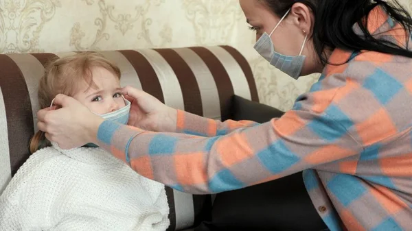 Anne çocuğuna nasıl tıbbi maske takılacağını gösterir. Annem küçük kızı için nasıl tıbbi maske takılacağını öğretiyor. Evde kal anne. Coronavirus veya COVID-19. Evde aile, viral hastalıkların önlenmesi — Stok fotoğraf