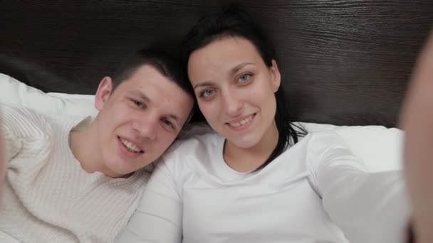 Ett ungt par som håller en smartphone i handen fotograferas tillsammans, filmar video blogg på sängen. Lycklig familj make och fru ta bilder på nätet med hjälp av en modern mobiltelefon hemma på soffan i sovrummet. — Stockvideo