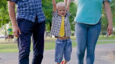 Baba, oğul, anne şehir parkında oynuyor, mutlu çocuk ebeveynlerin elini tutuyor, zıplıyor. Mutlu aile yürüyüşleri, parkta oyunlar bahar ve yaz aylarında el ele tutuşmalar. Takım çalışması. Sağlıklı çocukluk