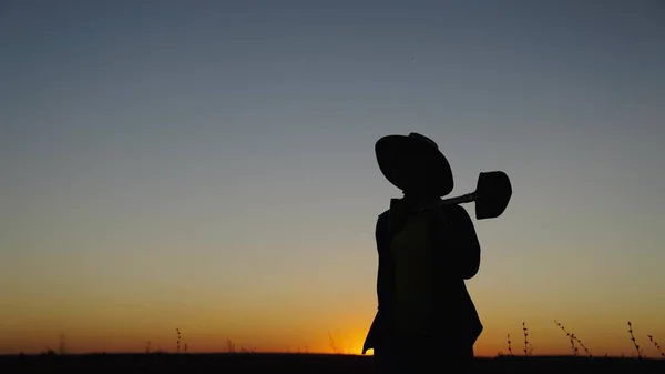 Silhouette A woman farmer with a shovel in her hands goes through field from work. A worker with a shovel walks through the plantation at sunset. Agricultural business, agriculture. Growing vegetables