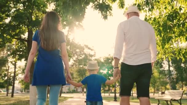 Feliz familia camina con un niño en la calle tranquila en verano al atardecer. Padres felices y niños caminando en el parque. Papá, hijo, mamá y tomad las manos en la acera en la ciudad bajo el sol. Vacaciones familiares, fin de semana — Vídeos de Stock