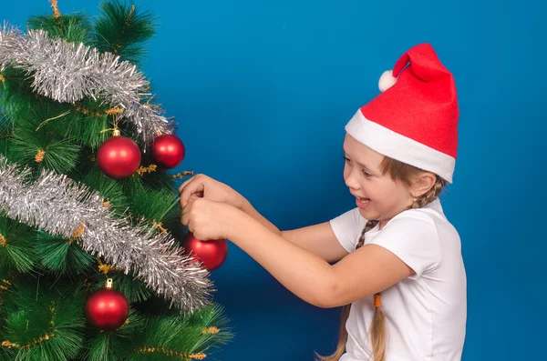 A menina em um gorro vermelho pendura em um abeto com ouropel um brinquedo um — Fotografia de Stock