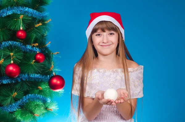 一顶红帽子头成本的女孩附近的一个优雅的新年树和掌控雪球微笑. — 图库照片