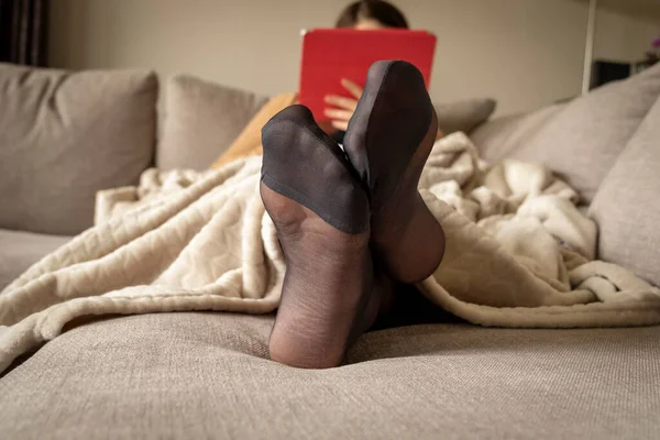 A portrait of a woman\'s feet in pantyhose while she is lying and resting in a couch under a warm and cosy fleece blanket reading and surfing on her tablet.