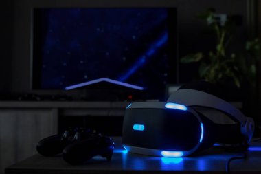 Brecht / Belçika - 12 Haziran 2019: Kalibrasyon ekranı ile arka tarafta televizyonu açık olan karanlık bir masada sanal bir playstation kulaklığı.