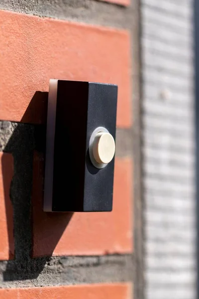 A close up portrait of a doorbell on a red brick wall. The device is next to a door and is black with a white button to be pressed to ring the doorbell.