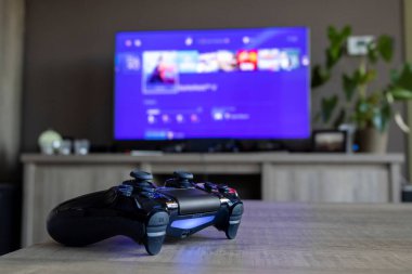 BRECHT, BELGIUM MARCH 28 2019 A wide portrait of a Sony Playstation 4 controller on a wooden table in front of a telivision showing the PS4 home screen in a living room. clipart