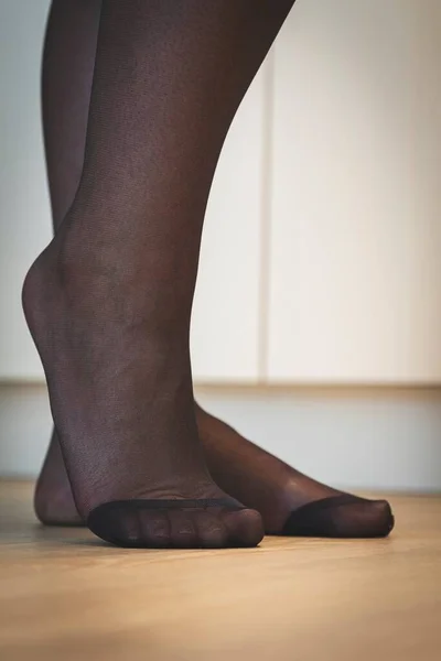 A close up portrait of the elegant feet of a girl in black pantyhose with the nylon at the toes being reinforced standing on a wooden floor and posing.