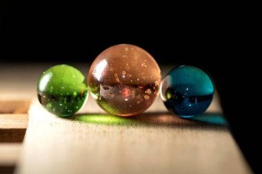 Ahşap bir yüzey üzerindeki üç farklı renkli bilyenin yakın plan portresi renkli yansımalar yapıyor. Cam küreler turuncu, yeşil ve mavi..